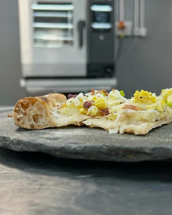 Pizza chiara servita su un tagliere di pietra, davanti al forno.